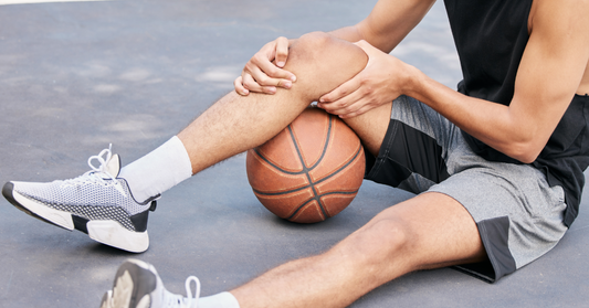 An image of a man sitting on a blacktop in workout attire with a basketball underneath his knee while he holds his knee in his hands emphasizing joint pain and slow workout recovery and how BPC-157 and GH-Release can help with recovery