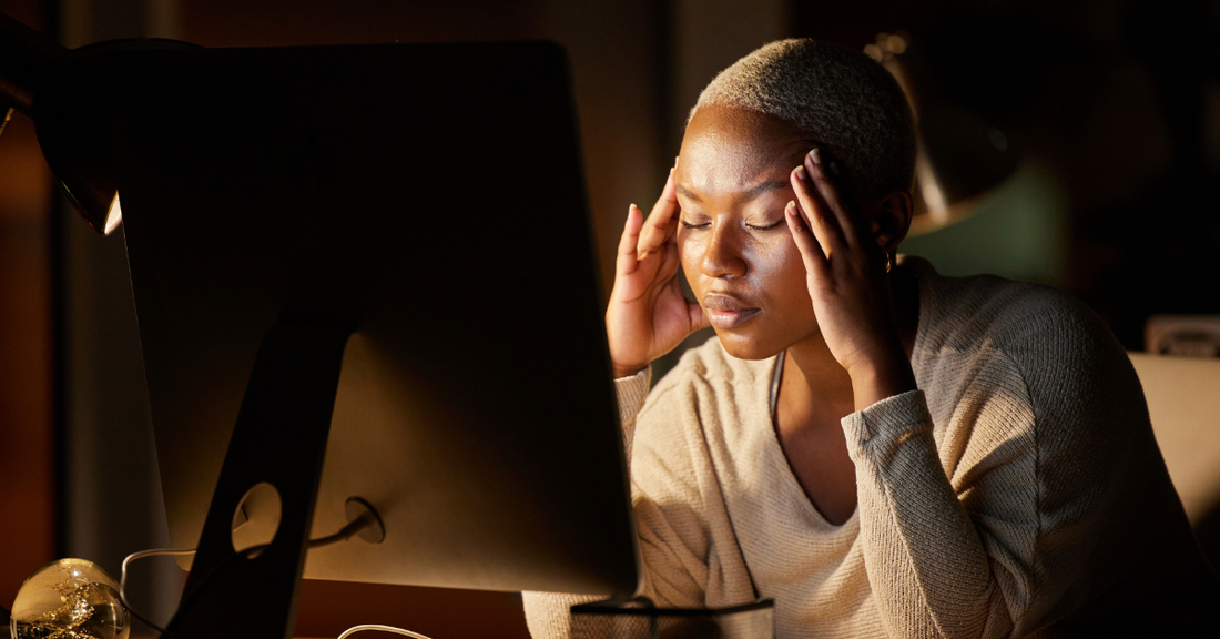 An image of a woman sitting at her computer in a dark room with her head in her hands and her eyes closed emphasizing how peptide nootropic capsules can help with brain fog and unmotivation